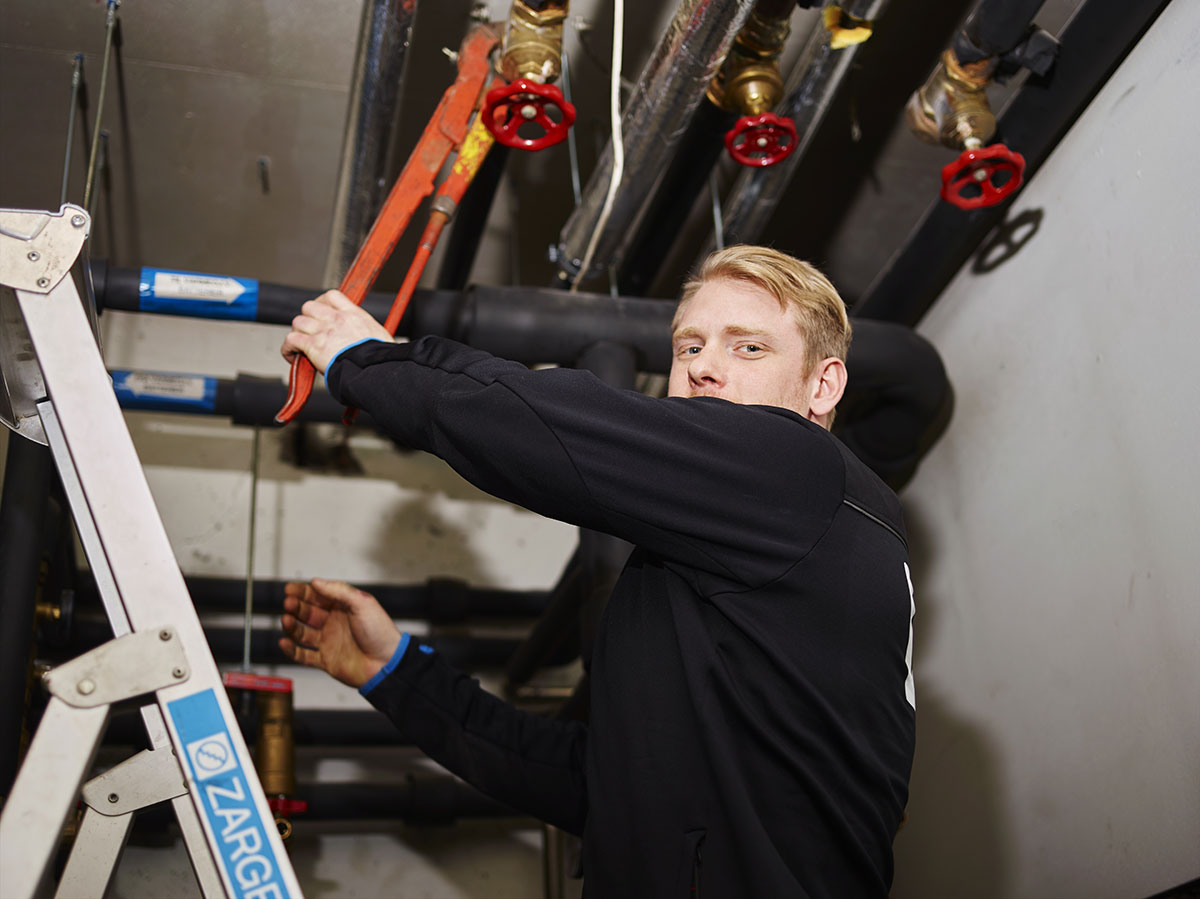 Rørlegger fra Kråkerøy Rør jobber med rørsystem i teknisk rom, bruker rørtang for å stramme koblinger på stoppekraner i himlingen.
